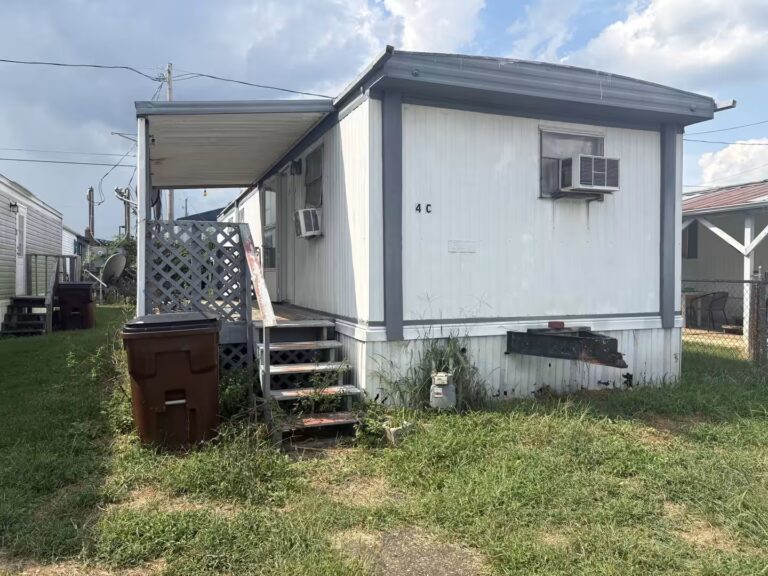 Weathered white mobile home marked 4C with wooden porch stairs and trash can in grassy yard