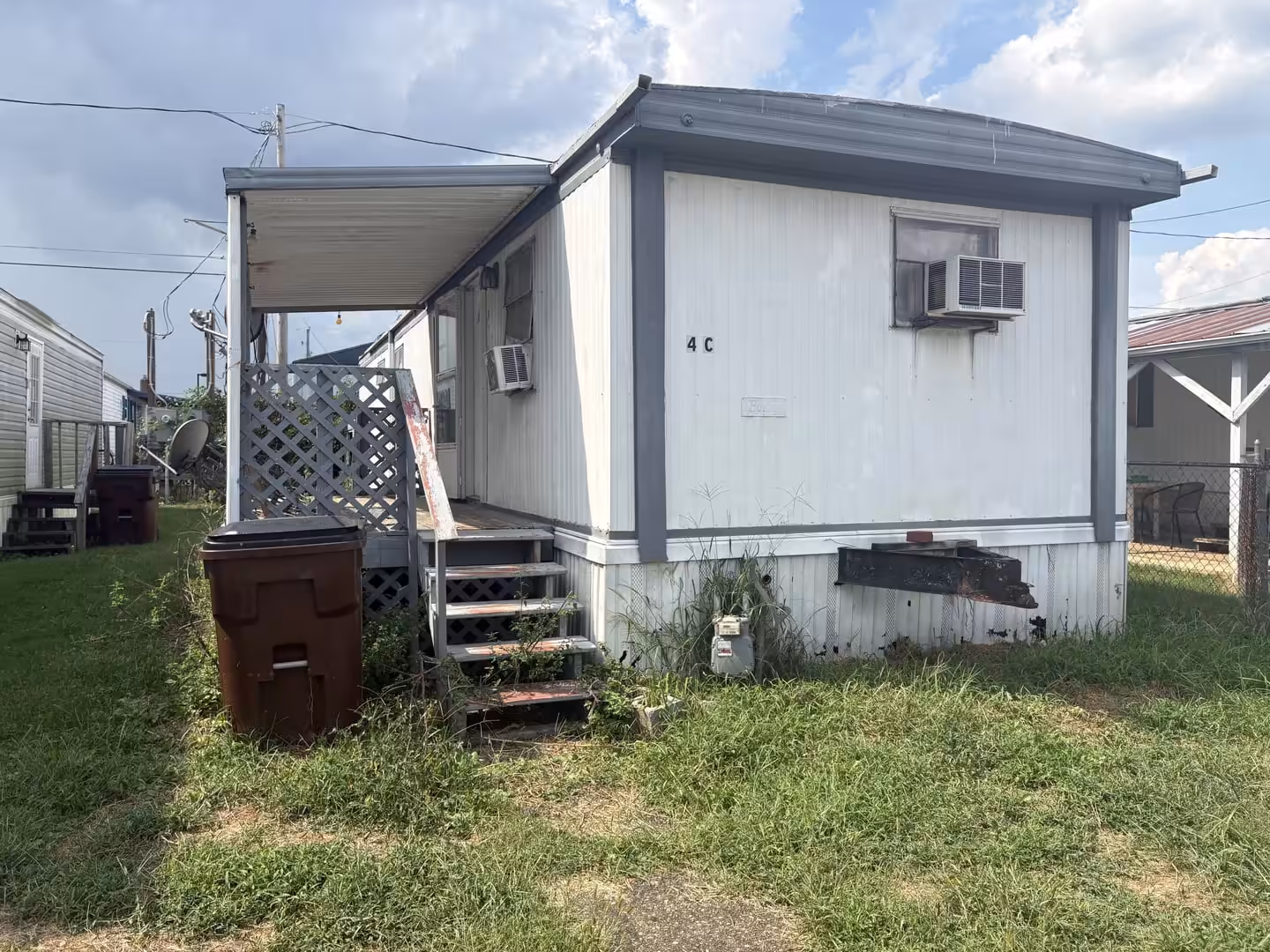 Weathered white mobile home marked 4C with wooden porch stairs and trash can in grassy yard