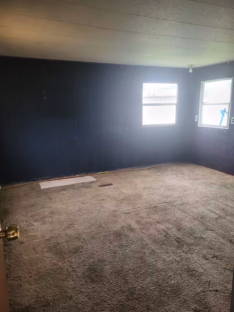 Empty room with dark blue walls, brown carpet, and two bright windows on the right wall.