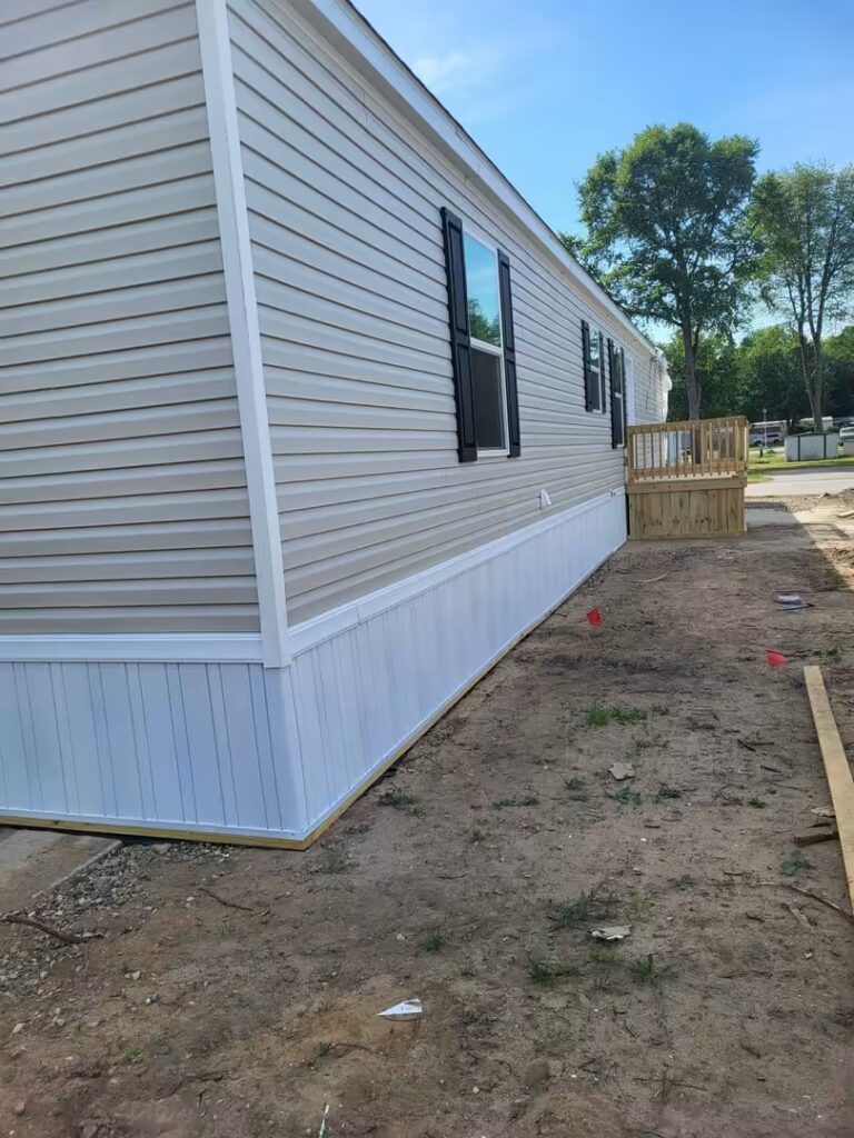 Mobile home with beige siding, black shutters, and small wooden deck beside dirt yard