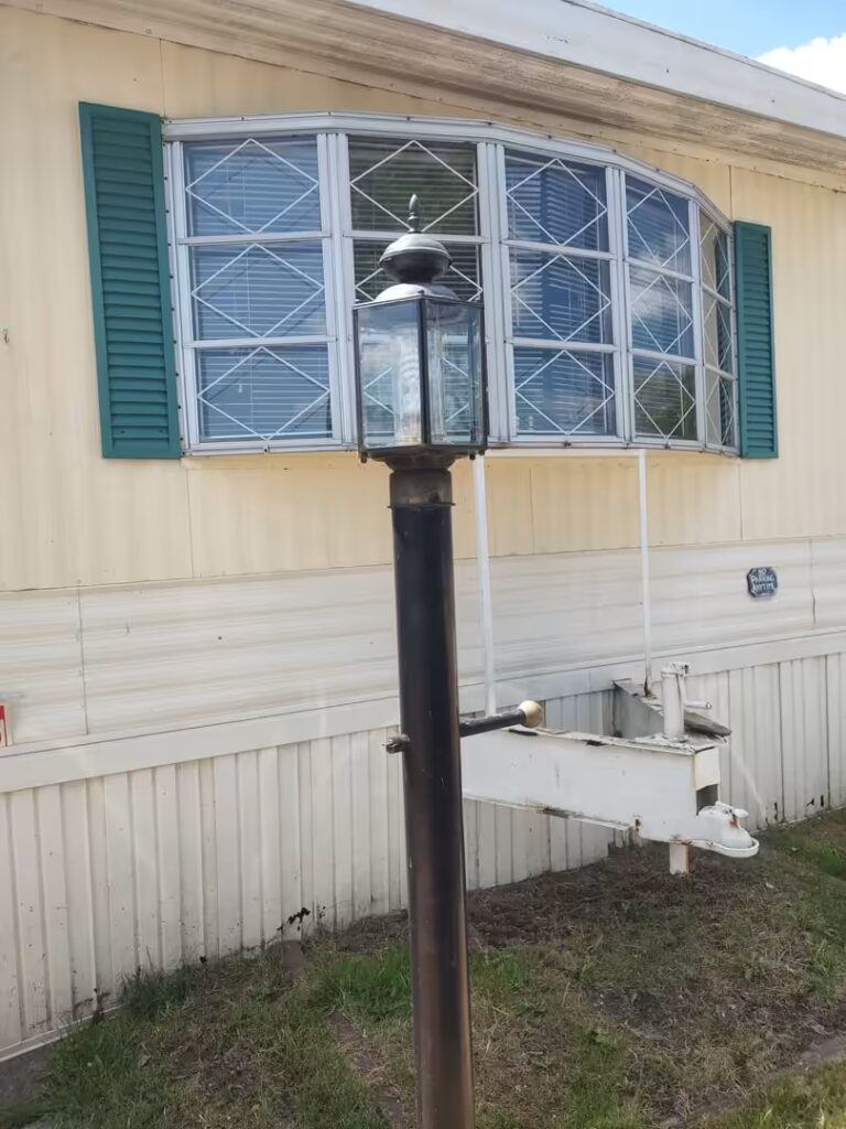 Black lamp post in front of a mobile home with a barred bay window and green shutters.