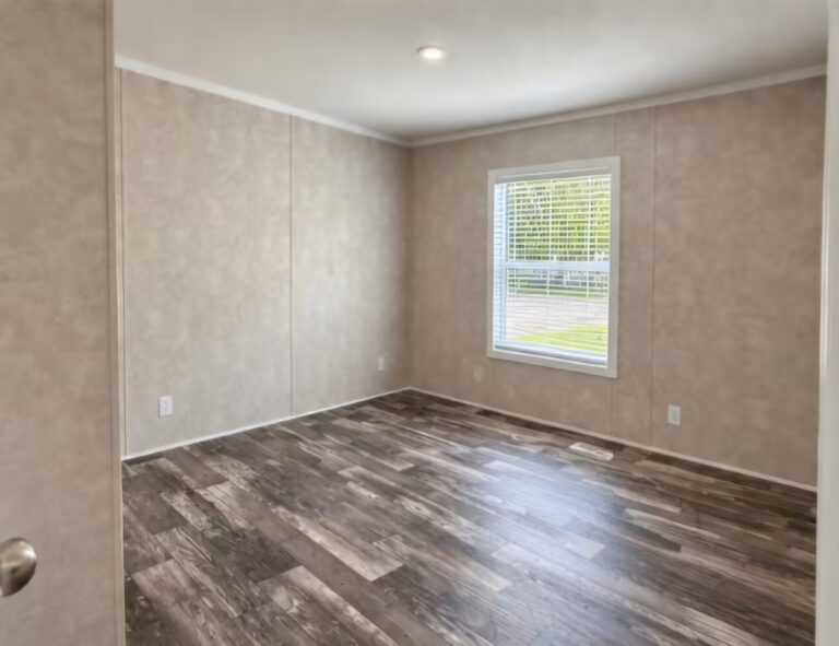 Empty room with beige walls, wood-look flooring, and a window with white blinds overlooking greenery.