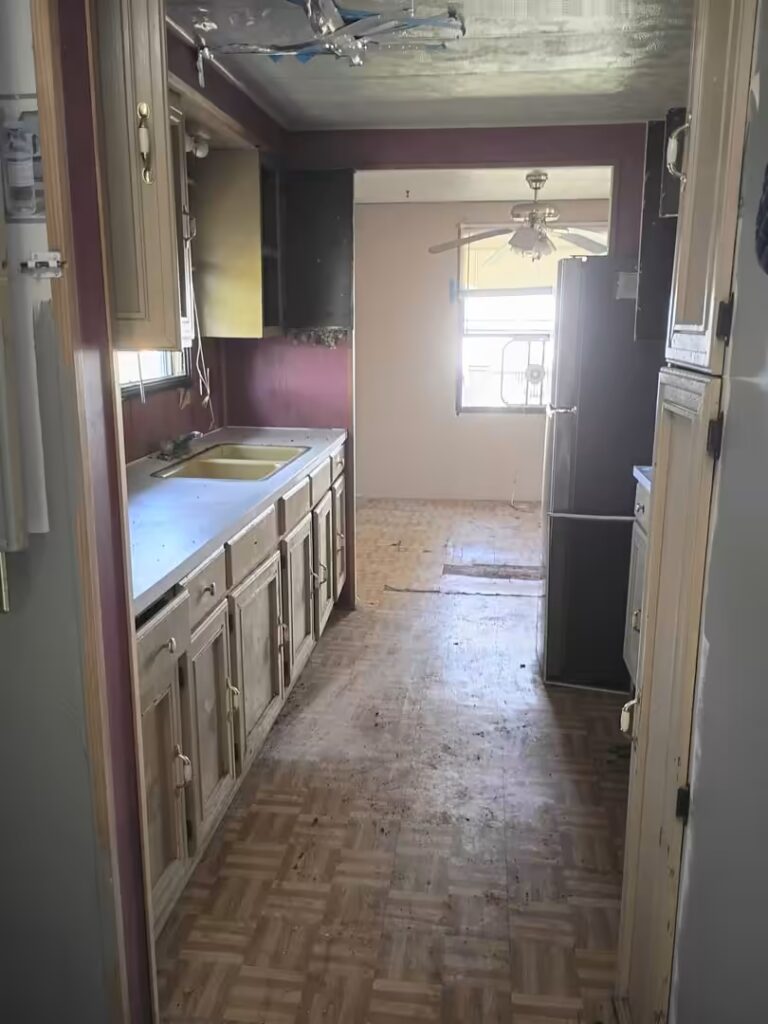 Dilapidated kitchen with damaged ceiling, old cabinets, worn parquet floor, and refrigerator