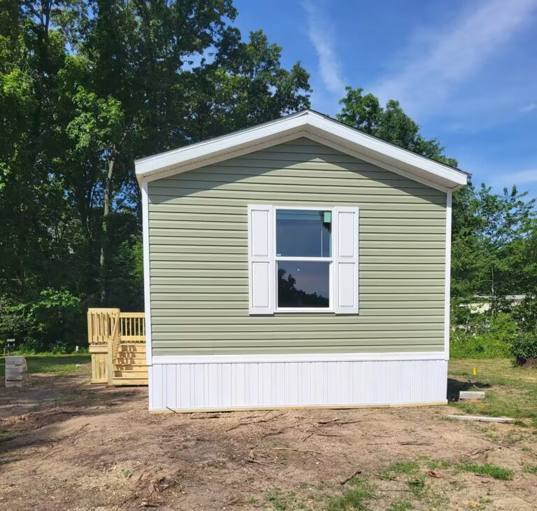 Small green house with white trim, shutters, and wooden deck stairs on a dirt lot surrounded by trees.