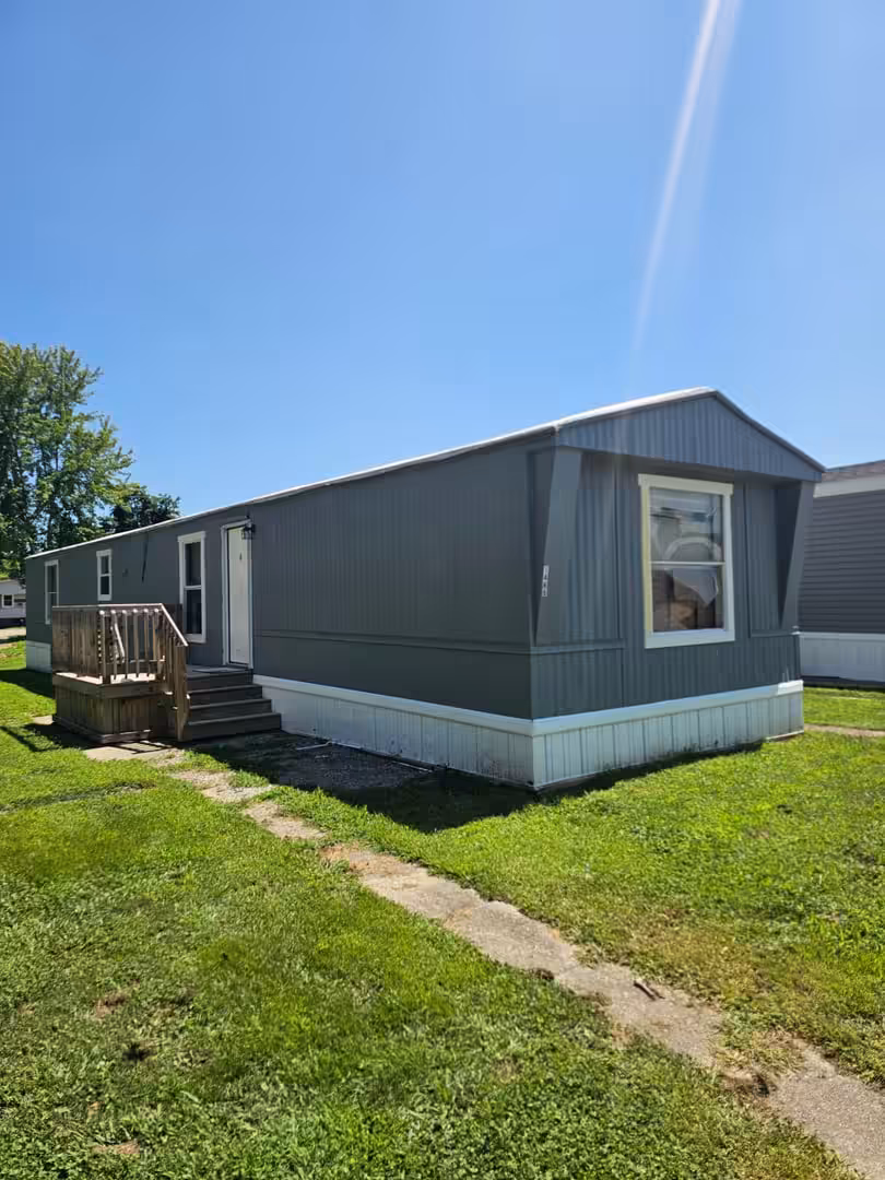 Gray single-wide mobile home with wooden deck and stairs on grassy lawn under blue sky.