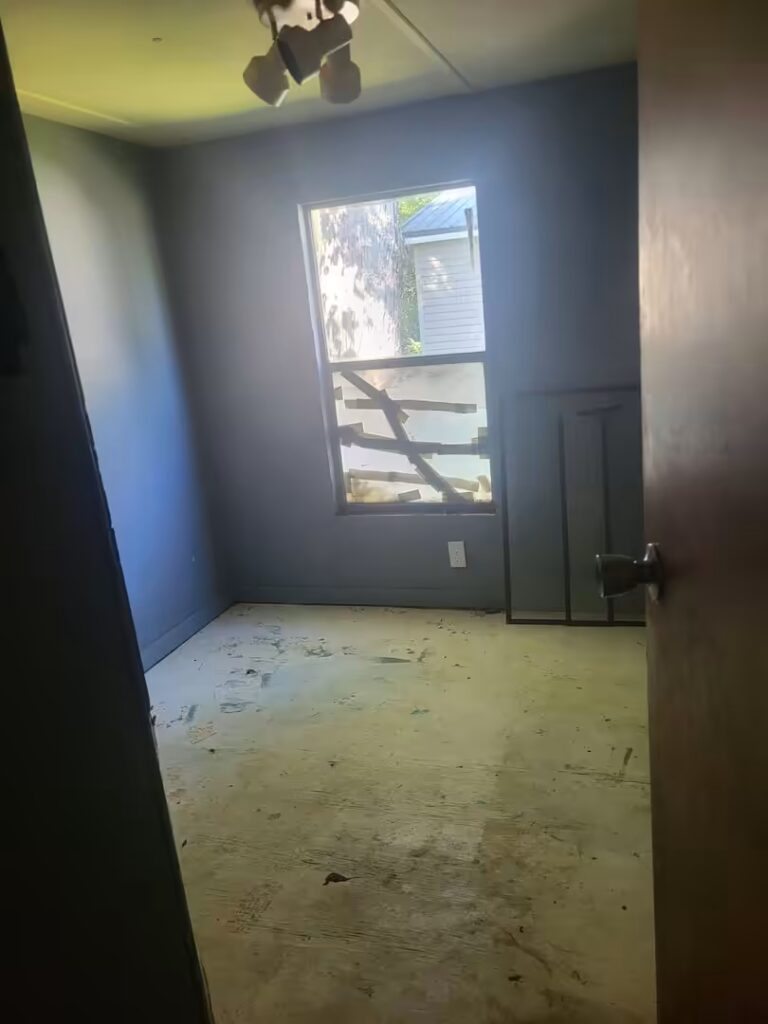 Empty room with broken boarded window, bare stained floor, blue-gray walls, and exposed ceiling light fixture.