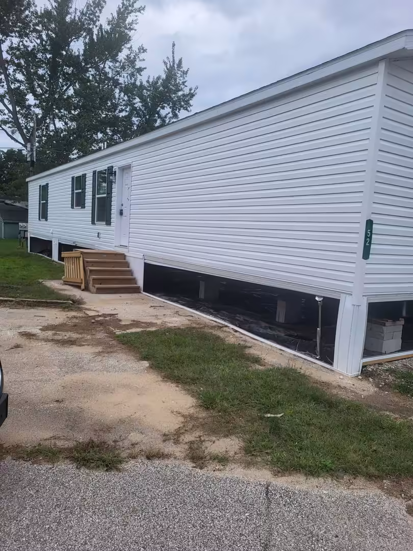 White mobile home with wooden entry stairs and elevated exposed foundation, lot number 52 on green marker