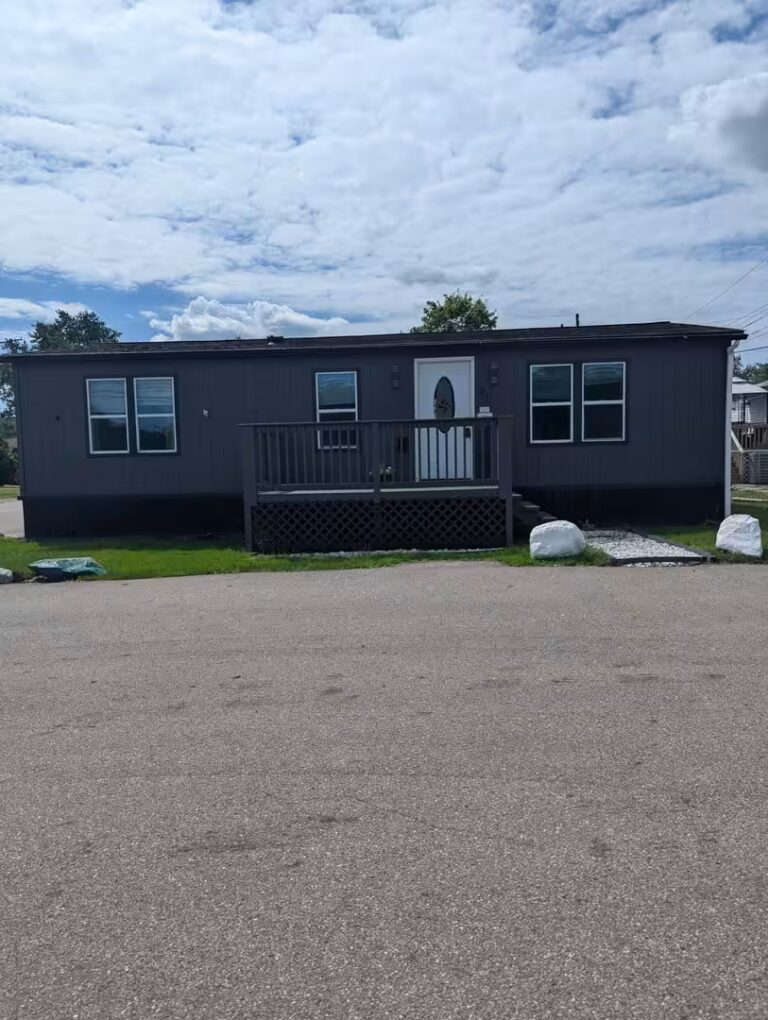 Dark gray mobile home with wooden front deck and white door under a cloudy sky.