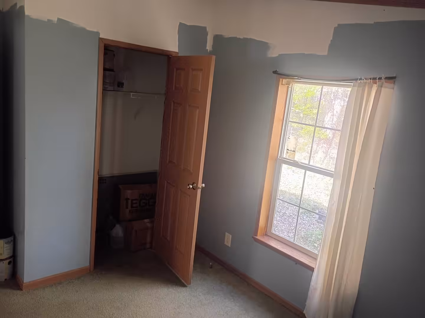 Room with partially painted blue walls, open closet door with TEGO box inside, and curtained window