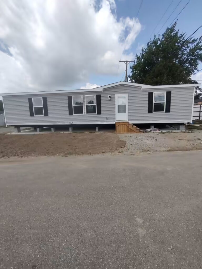 Gray mobile home with black shutters and wooden entry steps on a gravel lot under a cloudy sky.