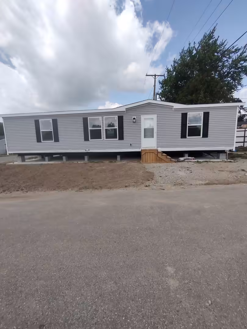 Gray mobile home with black shutters and wooden entry steps on a gravel lot under a cloudy sky.