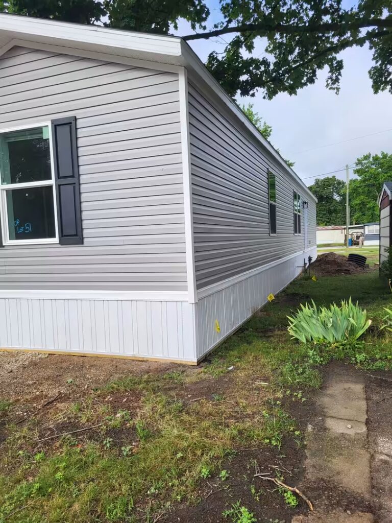 Gray mobile home with white skirting, black shutters, and "Lot 51" written on its window.