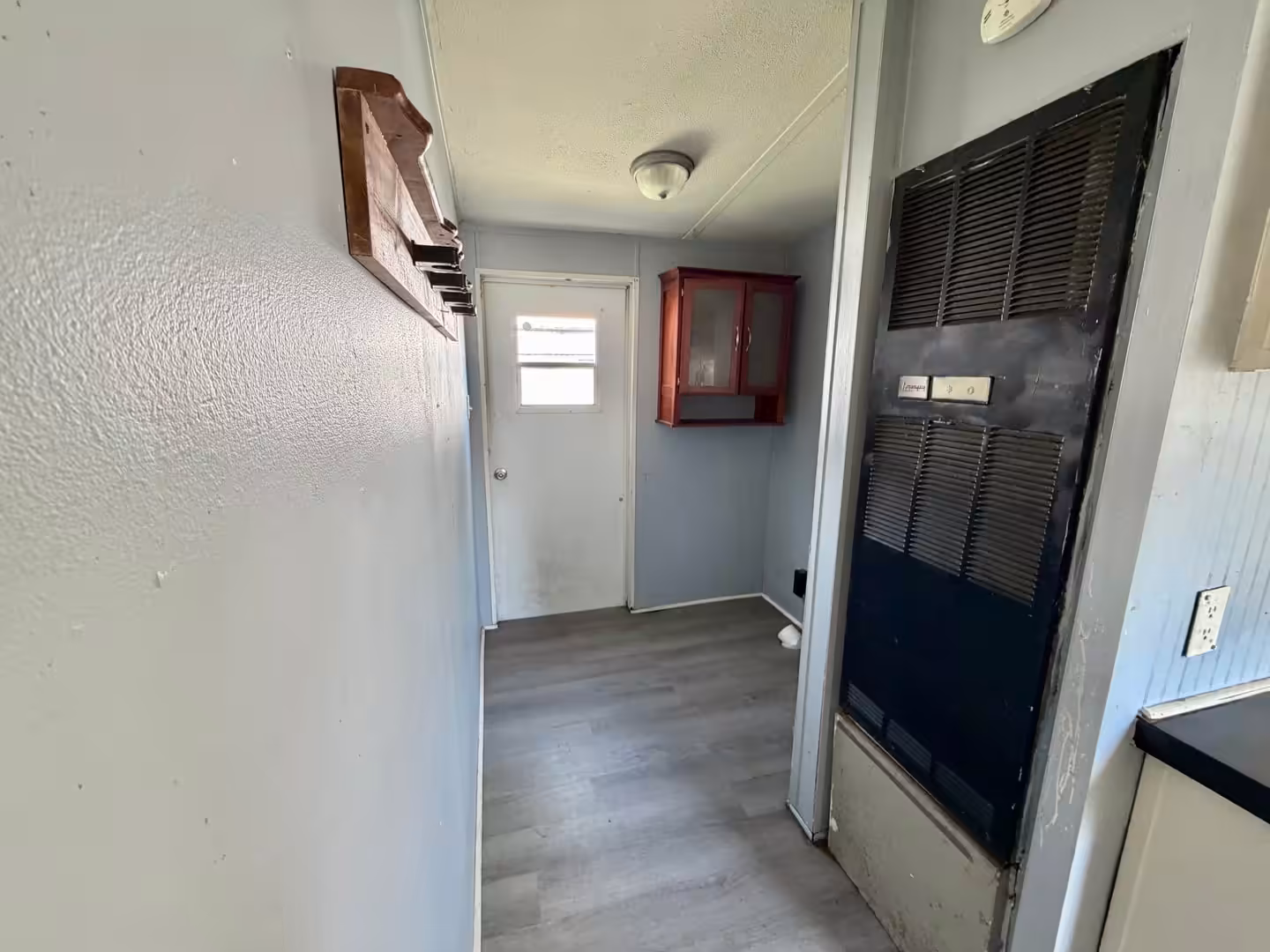 Narrow room with white door, red wall cabinet, and black wall furnace on wood-look flooring
