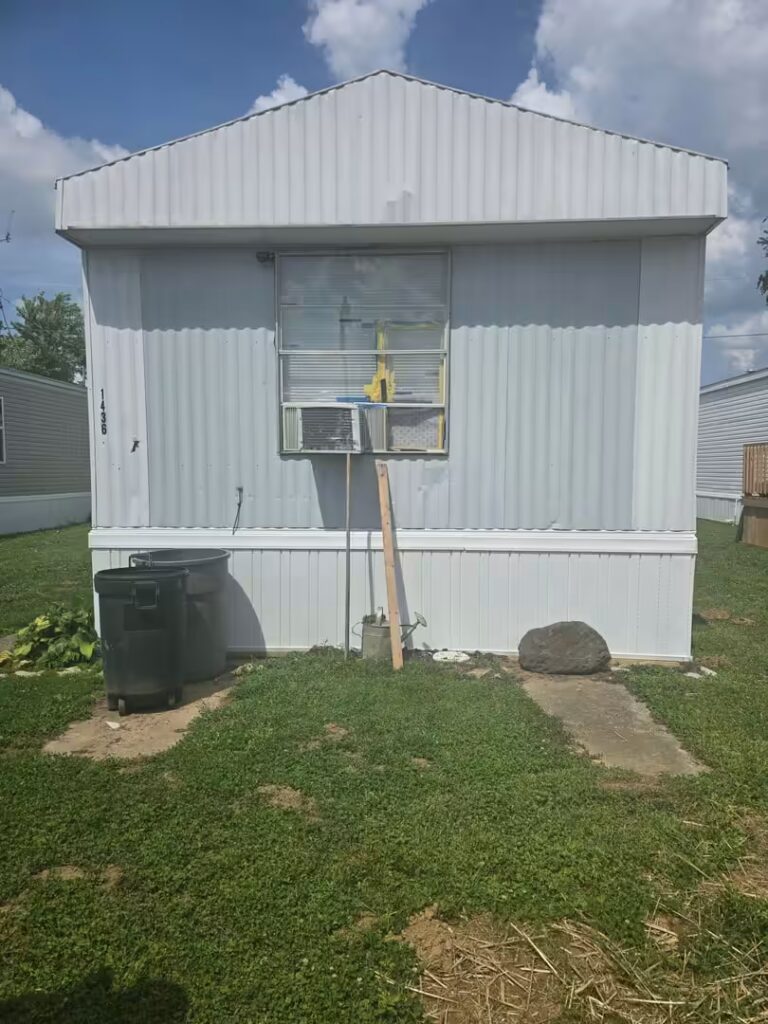 White mobile home with number 1436, window air conditioner, trash cans and board against siding.