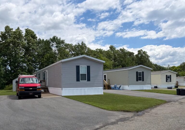 Red van parked beside a gray mobile home, with more mobile homes, lawn, and trees under a cloudy sky.